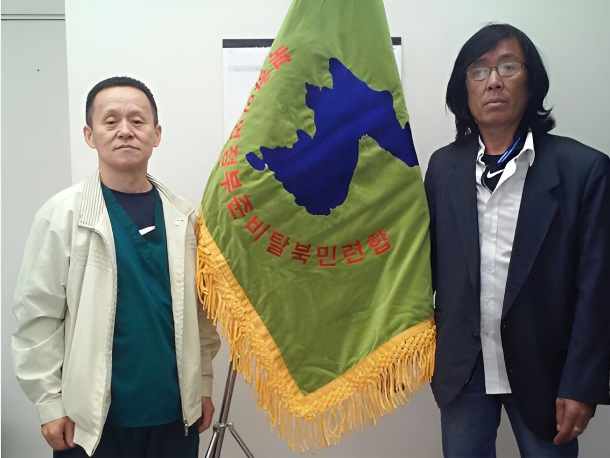 two men stand indoors beside a green flag featuring a blue map silhouette of korea and red korean text with golden fringe along the bottom edge