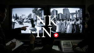 control room with two large crt screens showing a crowd in an urban setting bold nk in text with a red star overlayed in the center