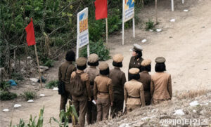 Group of women in brown military-style uniforms listening to a woman in a cap outdoors near signboards with Korean text by a dirt path in a rural area