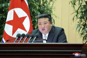 Kim Jong Un speaking at a wooden podium with multiple microphones, North Korean flag in the background.