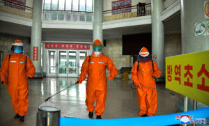 three workers in bright orange hazmat suits disinfect a spacious indoor lobby with spray wists wearing face shields and gloves