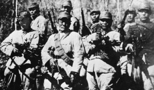 Black-and-white photo of soldiers in early 20th-century uniforms with rifles in a forest setting.