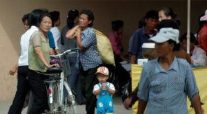 Toddlers blue overalls and striped cap stands in busy outdoor market, holding a cup.