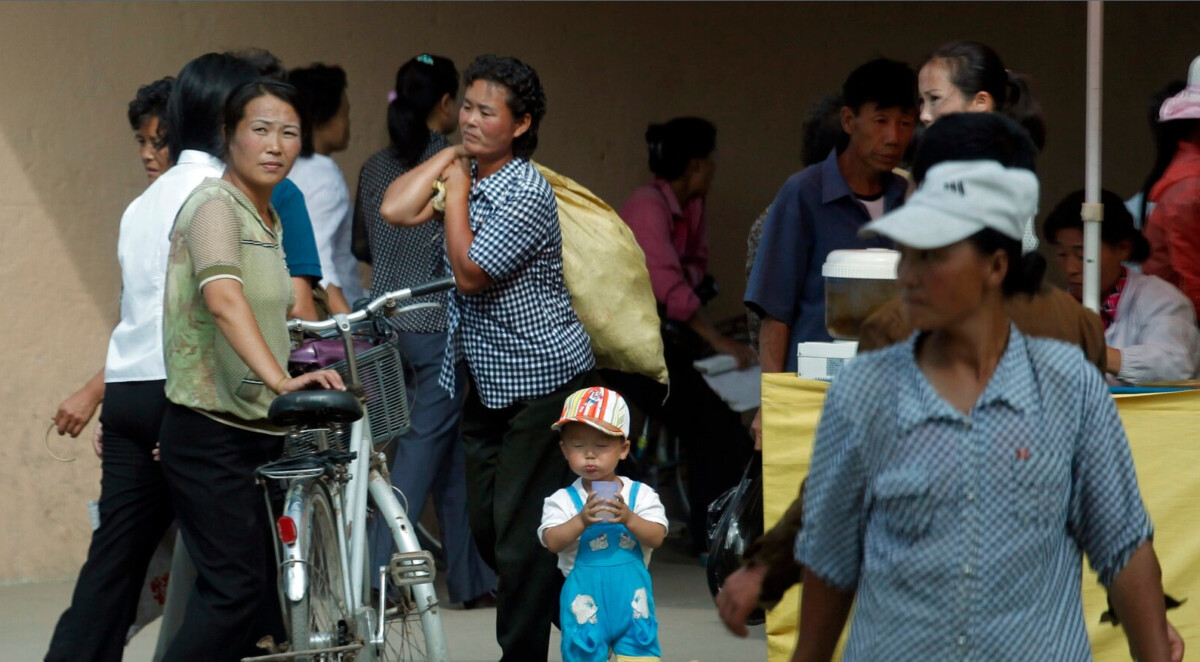 toddlers blue overalls and striped cap stands in busy outdoor market holding a cup