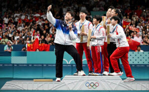 athlete in blue and white jacket takes a selfie on a podium with teammates holding awards olympic rings visible on the podium
