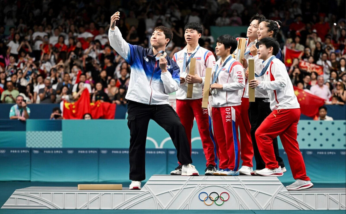 athlete in blue and white jacket takes a selfie on a podium with teammates holding awards olympic rings visible on the podium