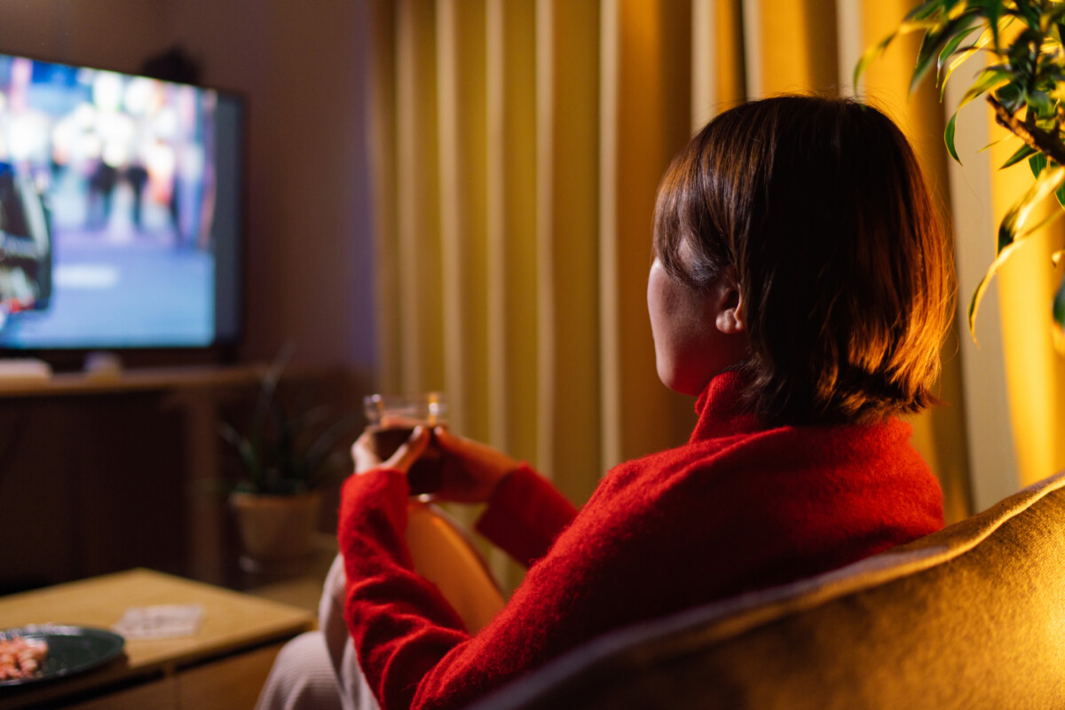 Rear view of young woman watching movie at home at night | nk insider person with short dark hair in a red sweater sits on a couch watching tv while holding a glass