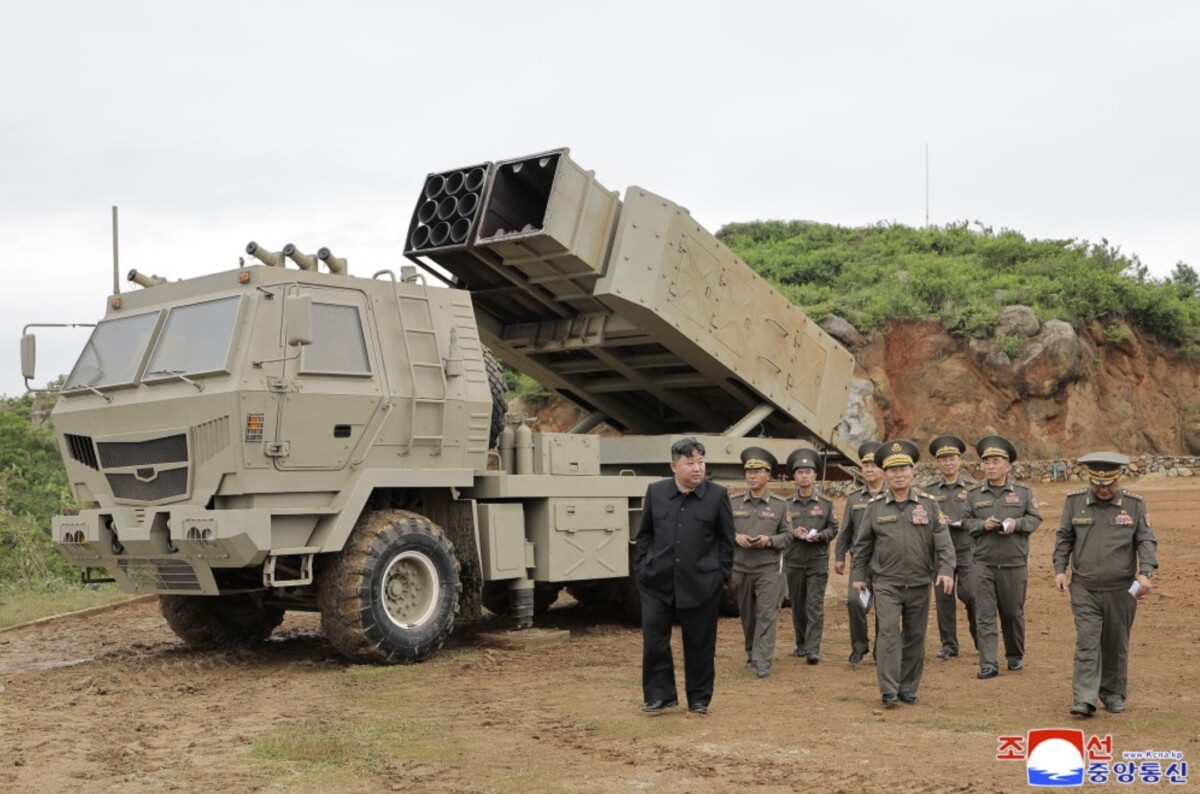 Kju at a missile test site 1 | nk insider a man in a black suit walks beside a line of uniformed officers next to a large beige rocket launcher mounted on a heavy truck in a dirt area with a rocky hillside behind