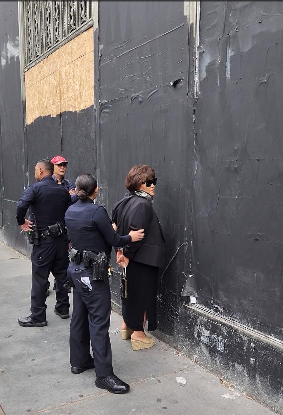 three police officers confront a woman in black outside a tall dark wall on a city sidewalk the woman wears sunglasses and beige wedge shoes