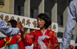 woman in a red floral hanbok joins hands with a partner during an outdoor traditional dance in front of a building