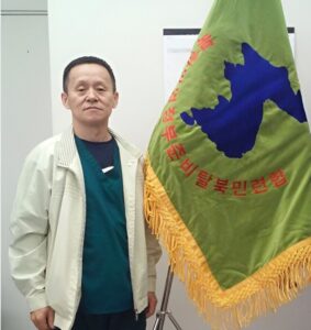 Two men stand indoors beside a green flag featuring a blue map silhouette of Korea and red Korean text, with golden fringe along the bottom edge.