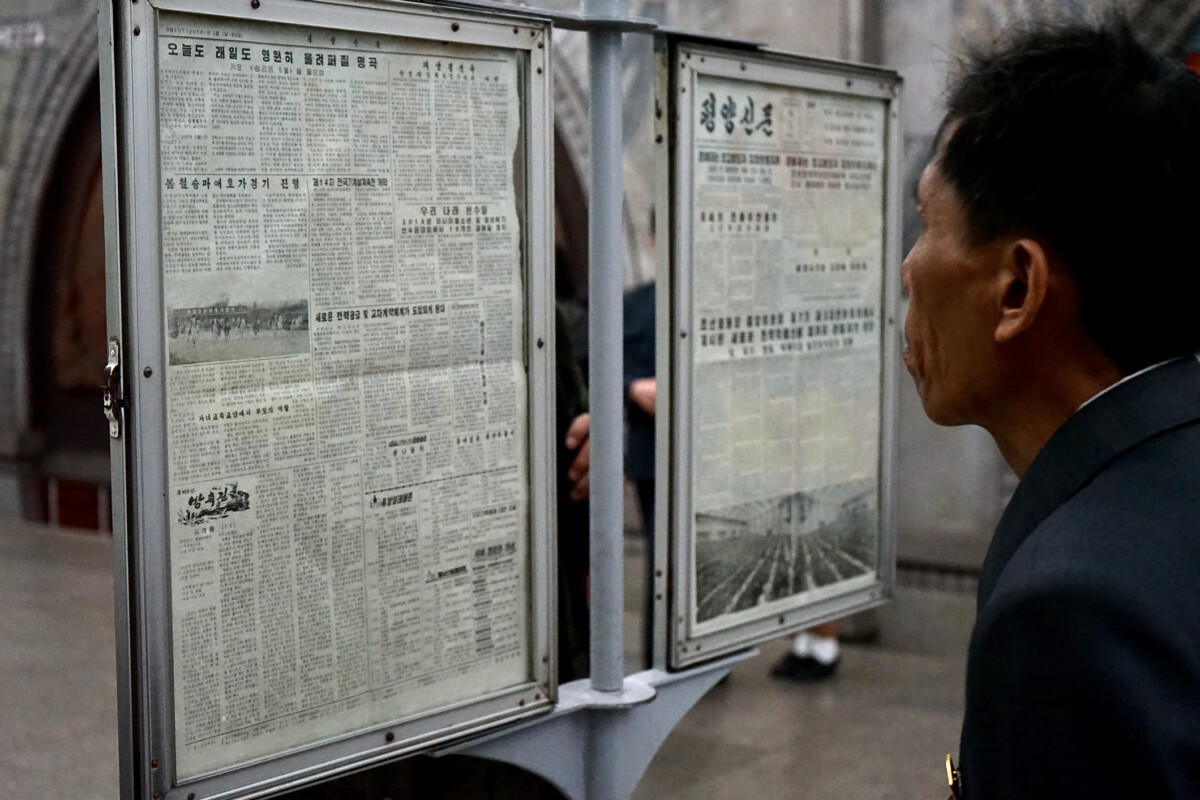 Korean news | nk insider side profile of a man in a suit reading korean newspaper pages mounted in glass display cases outdoors