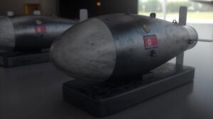 two gray cylindrical bombs on dark wooden bases sit on a smooth display table in a dim room with small flag stickers on their sides