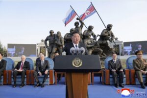 a man in a dark suit giving a speech at a podium with multiple microphones during an outdoor ceremony with a large bronze statue and flags in the background