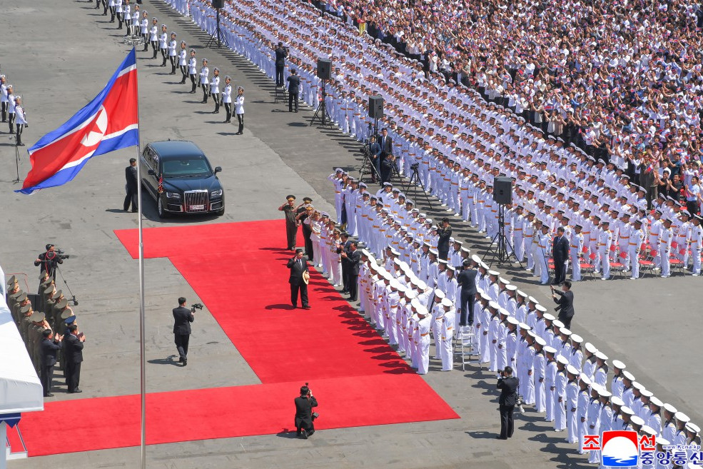 north korean flag beside a red carpet parade of uniformed cadets officials photographers and a large crowd watch from the stands