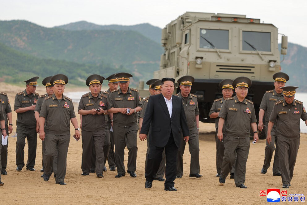 group of uniformed military officers walking on a sandy beach with a large armored vehicle parked behind them