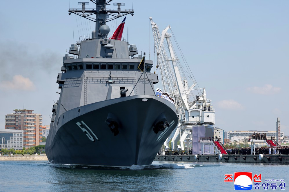 bow view of a gray military destroyer docking at a harbor with large white cranes and people on the pier in the background
