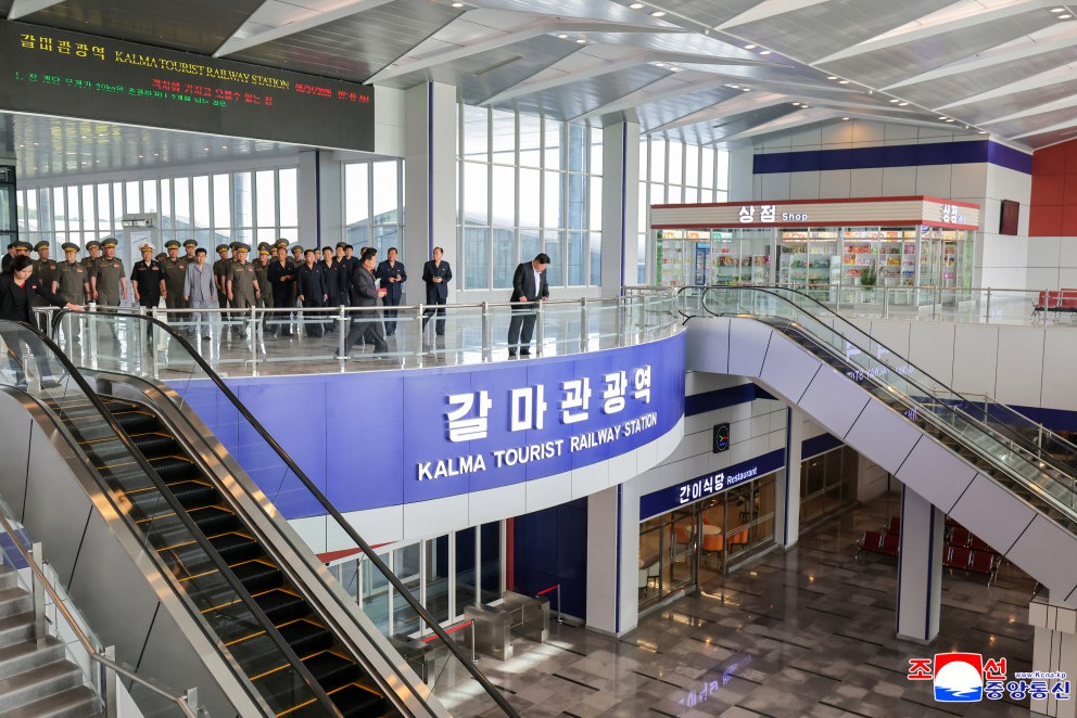 group of uniformed officials and civilians lined up on the upper level of kalma tourist railway station beside a blue sign reading kalma tourist railway station