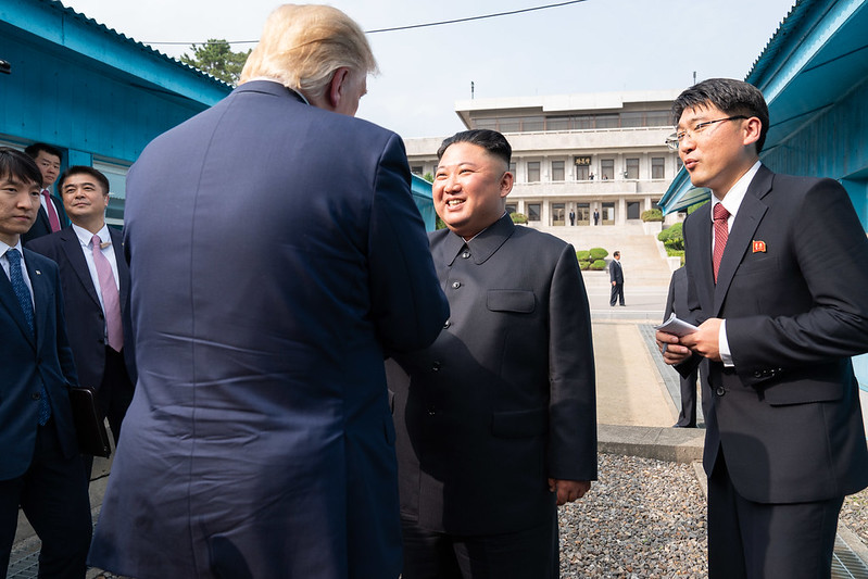 two world leaders shake hands in a formal outdoor meeting surrounded by aides in suits and a government building in the background