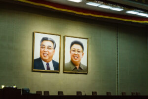 two framed portraits of two smiling men hanging on a green wall above a row of chairs and a stage with microphones and desks below