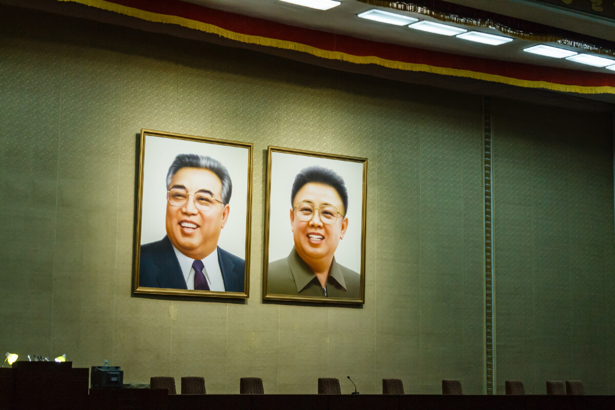 two framed portraits of two smiling men hanging on a green wall above a row of chairs and a stage with microphones and desks below