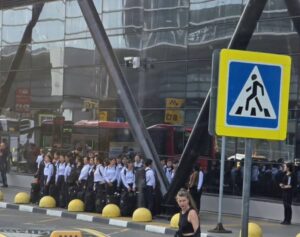 line of uniformed people with rolling suitcases waiting outside a glass front building a woman in a black top walks in the foreground near a pedestrian crossing sign