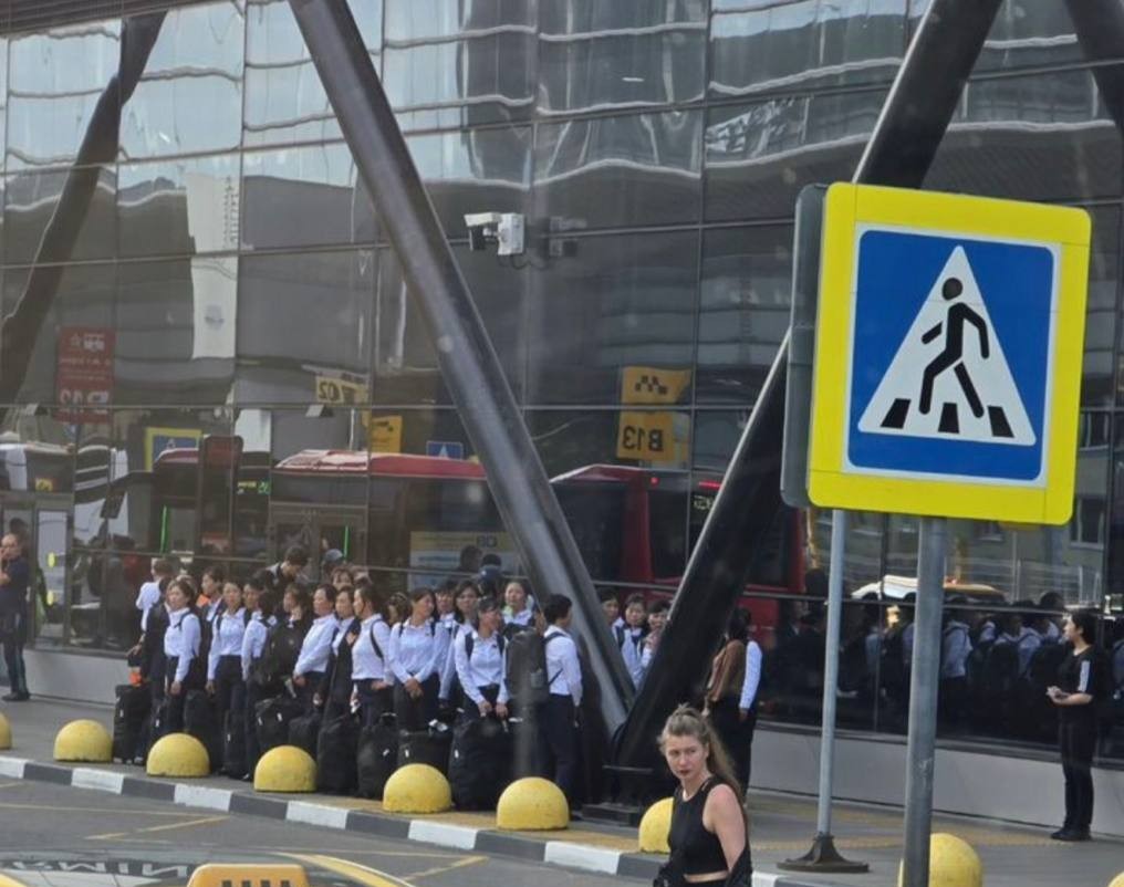 Photo20260817155535 | nk insider line of uniformed people with rolling suitcases waiting outside a glass front building a woman in a black top walks in the foreground near a pedestrian crossing sign