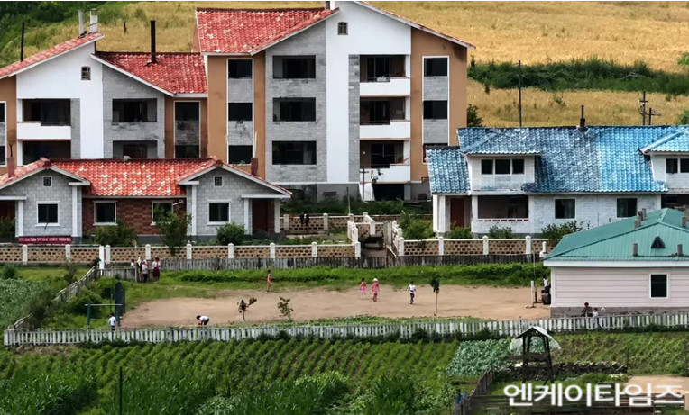 Village in nk | nk insider a row of newly built multi story houses with red and blue roofs behind a fenced dirt playground where children are playing with green fields and farmland in the foreground