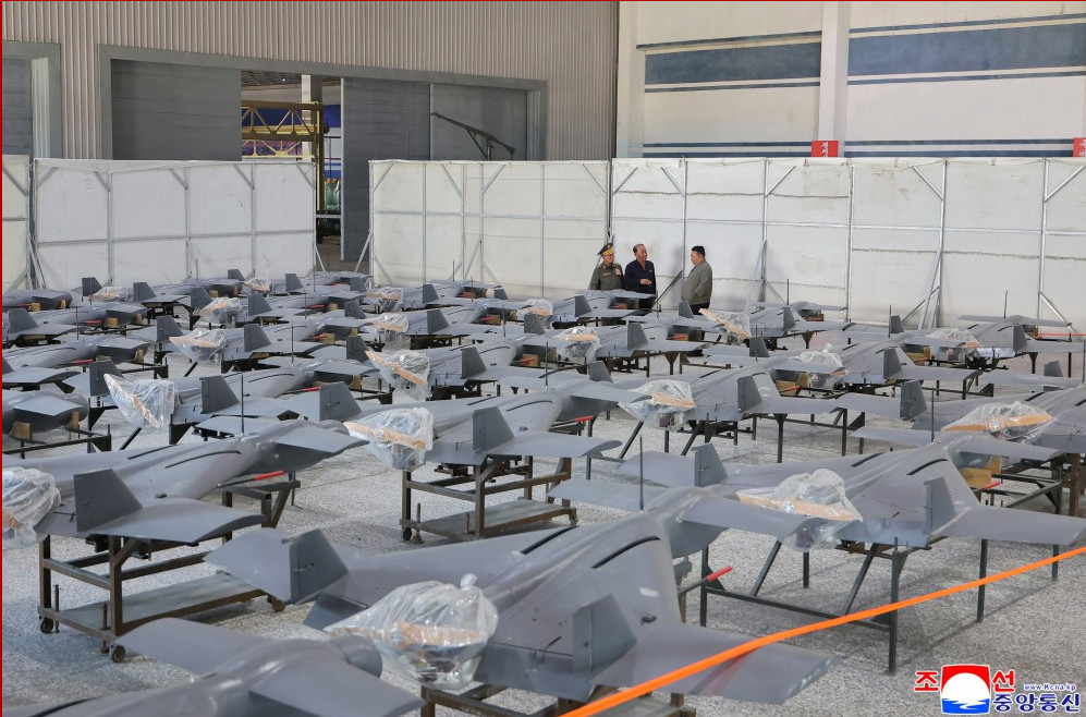 12 | nk insider row of gray unmanned aircraft fuselages on stands inside a hangar with three men talking in the background