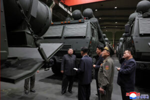 officials in dark suits and a uniformed officer inspect a row of large military armored vehicles inside a hangar