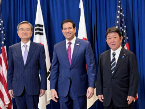 three men in suits stand side by side at a formal event with flags and a blue backdrop behind them