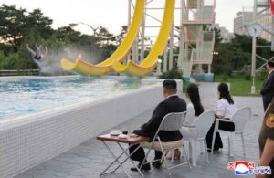 poolside spectators in formal attire sit on plastic chairs and observe a rider splashing into the pool from a large yellow water slide a small table with drinks is nearby