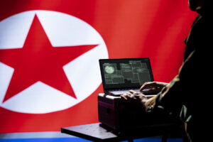 person typing on a laptop at a desk with a large north korea flag in the background
