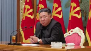 a man in a black suit speaks at a wooden desk with two microphones red flags in the background