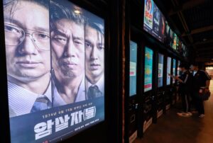 row of digital poster kiosks in a cinema lobby with a large three panel poster of a korean film showing three mens faces in the foreground