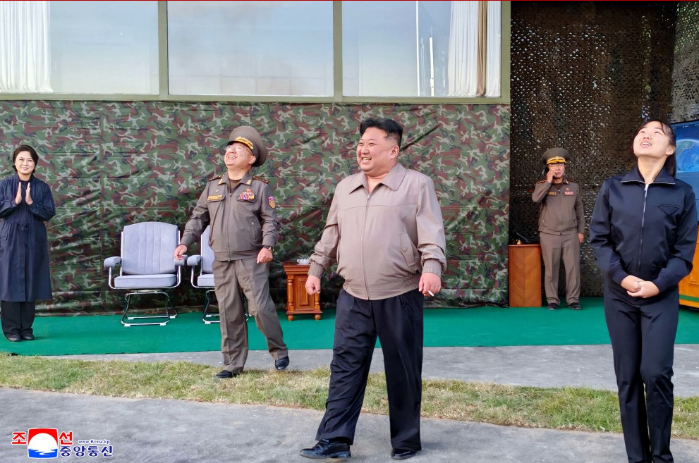 Kjukjalsj | nk insider group of officials and civilians standing outdoors on a stage with a camo backdrop looking up and smiling as if reacting to something overhead