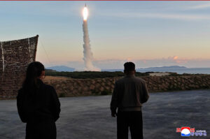 two people stand on a paved area watching a rocket launch from the coastline at sunset smoke plume rises as the rocket ascends over the sea