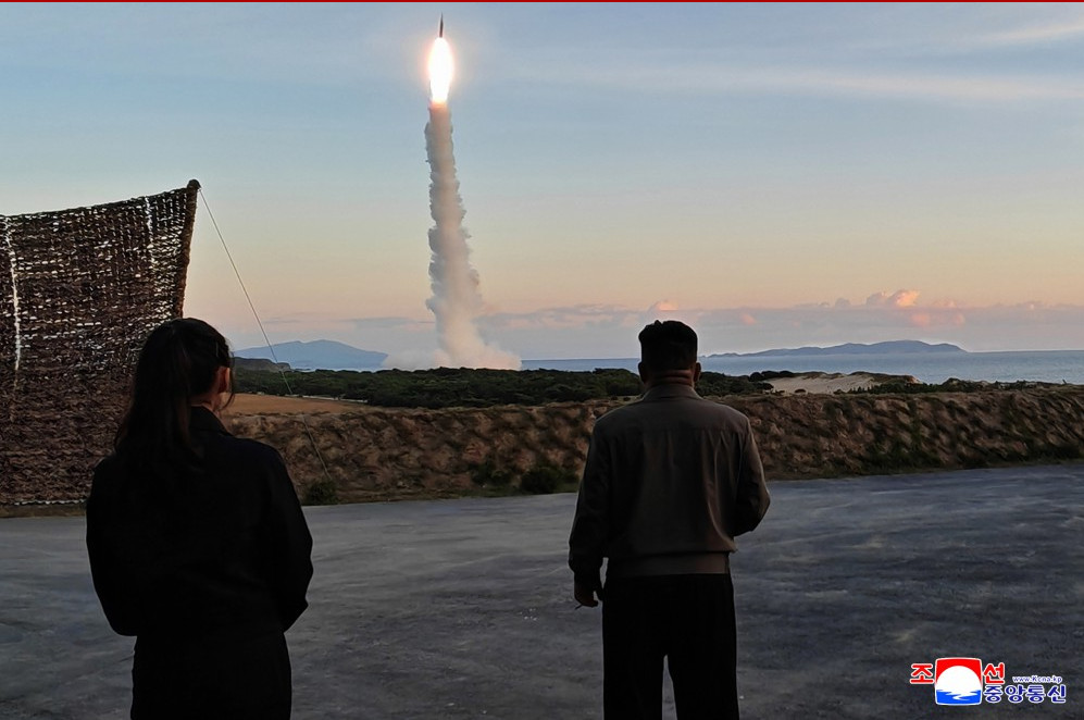 Kjumissletest | nk insider two people stand on a paved area watching a rocket launch from the coastline at sunset smoke plume rises as the rocket ascends over the sea