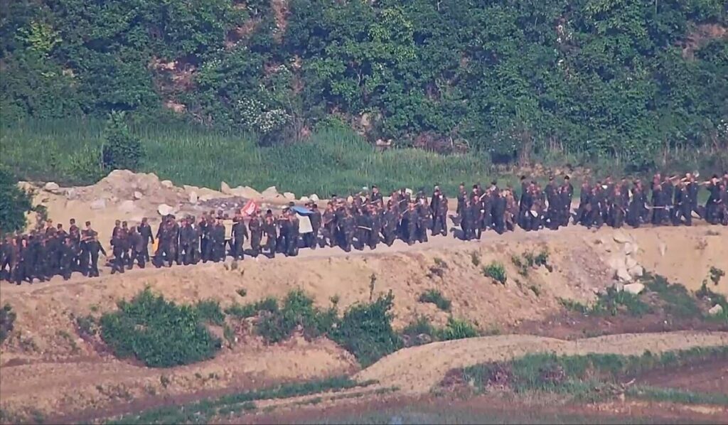 line of uniformed people marching in single file along a dirt road beside a grassy embankment in a rural area some banners visible